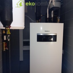 EKO KOMFORT - Biały piec grzewczy marki Buderus zamontowany na ścianie wyłożonej szarymi płytkami, z widocznymi rurami i zaworami, obok logo firmy EkoKomfort i fragment surowej ściany.