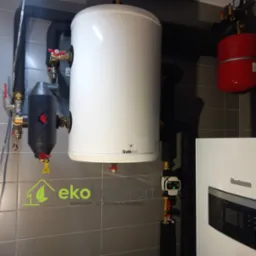 Biały zasobnik ciepłej wody Galmet z czerwonymi zaworami, czarny filtr, czerwony zbiornik wyrównawczy i piec Buderus na tle szarej ściany z płytek, widoczne rury instalacji centralnego ogrzewania...