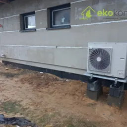 Zewnętrzna jednostka pompy ciepła na betonowych bloczkach przed ocieplonym budynkiem z widocznym logo firmy Eko Komfort na elewacji.