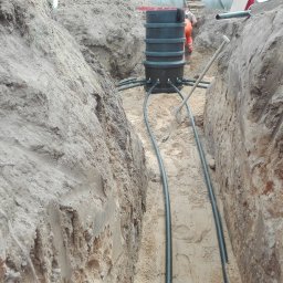 EKO KOMFORT - Wykop z piaskowym dnem, w którym umieszczono czarny, cylindryczny zbiornik z podłączonymi rurami; w tle widoczna osoba w pomarańczowym kombinezonie i łopata oparta o zbiornik.