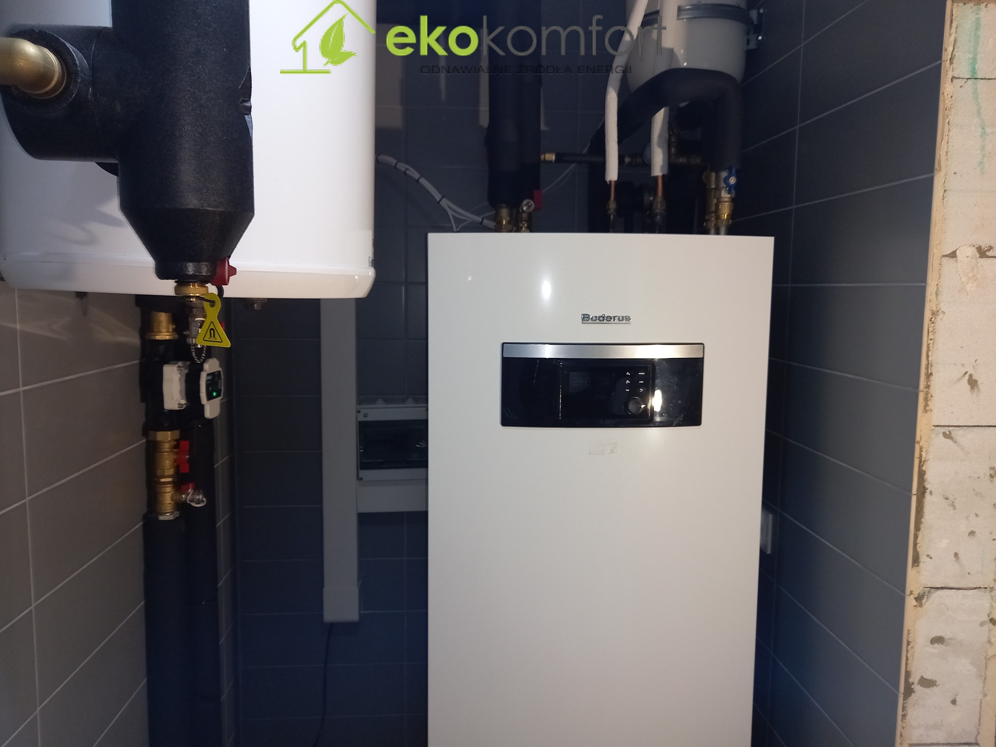 Biały piec grzewczy marki Buderus zamontowany na ścianie wyłożonej szarymi płytkami, z widocznymi rurami i zaworami, obok logo firmy EkoKomfort i fragment surowej ściany.