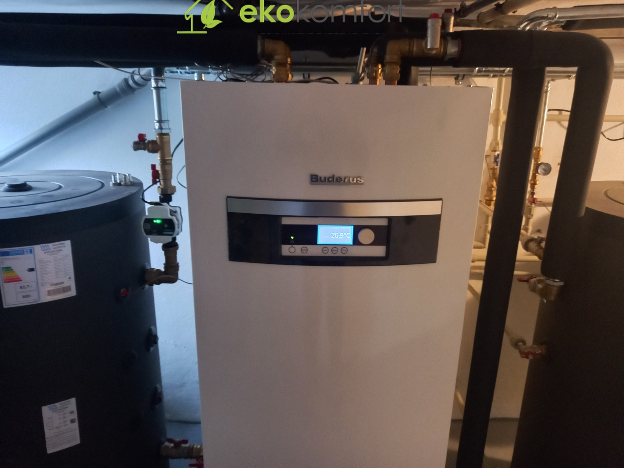 Biały piec grzewczy Buderus z wyświetlaczem 26.9°C, obok czarny zbiornik z etykietą energetyczną, widoczne rury i zawory w kotłowni. U góry logo firmy Eko Komfort.