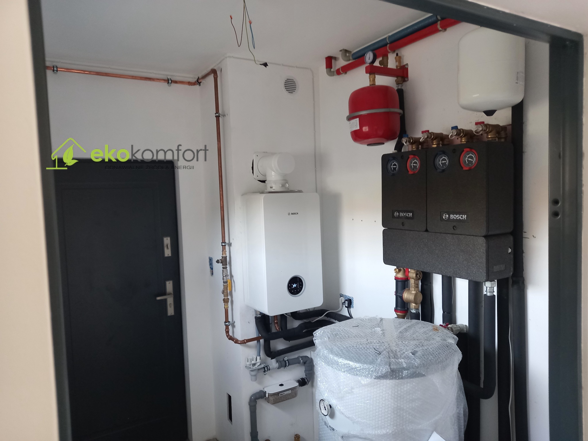 Nowoczesna kotłownia w jasnym pomieszczeniu z piecem gazowym Bosch, zbiornikiem buforowym owiniętym folią bąbelkową, instalacją hydrauliczną z czerwonym naczyniem przeponowym, oraz logo firmy Eko...