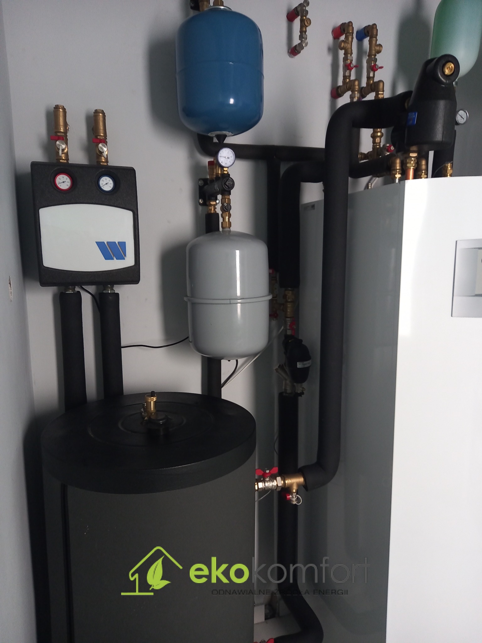 Kompletna instalacja grzewcza z kilkoma zbiornikami, czarnymi rurami izolowanymi termicznie i zaworami z czerwonymi pokrętłami, widoczny panel sterowania z manometrami i logo firmy Eko Komfort...