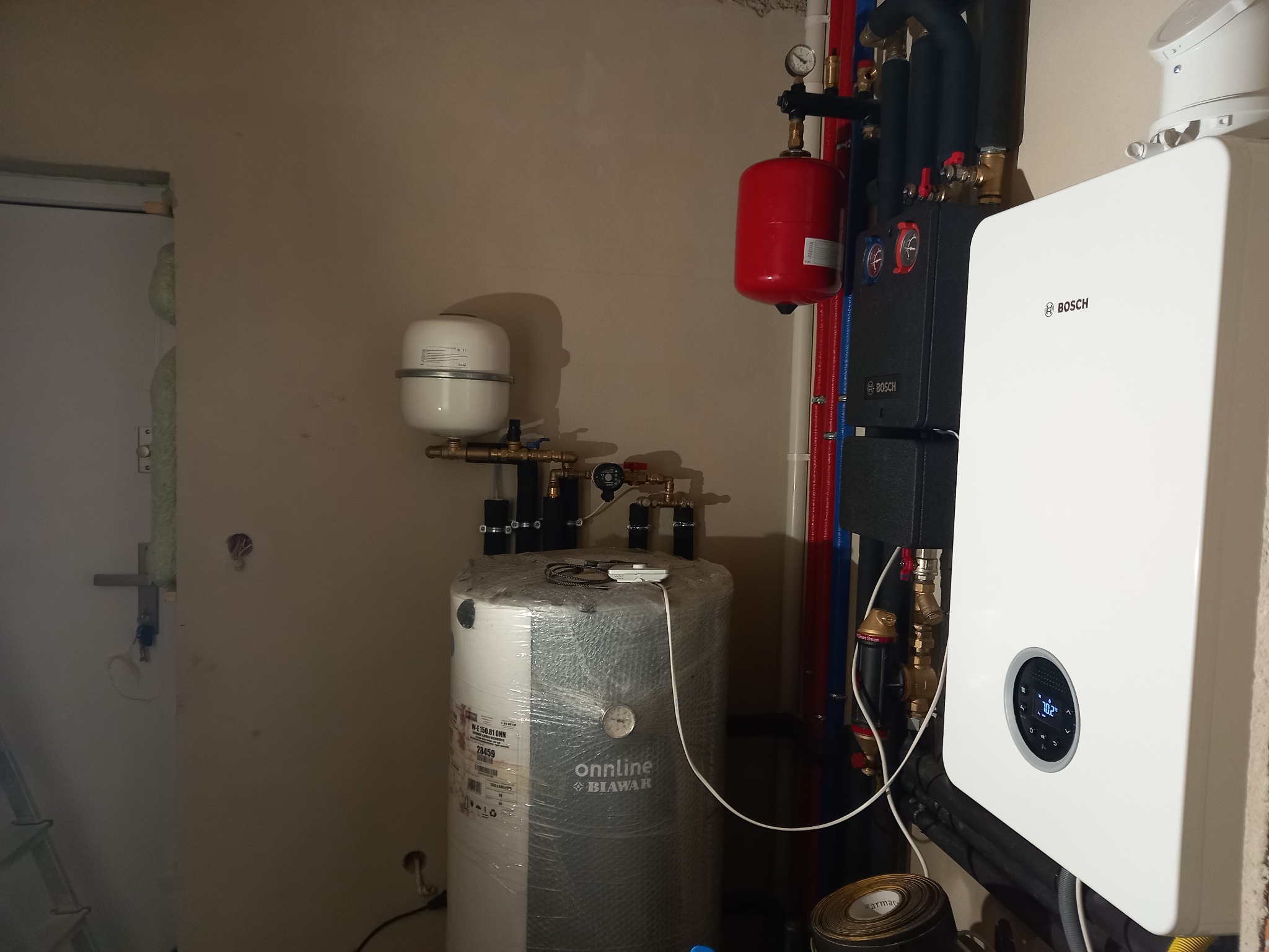 Biały piec gazowy Bosch z wyświetlaczem, podłączony do instalacji z czerwonym zbiornikiem wyrównawczym, obok owinięty folią ochronną zasobnik ciepłej wody użytkowej marki Onnline Biawar.