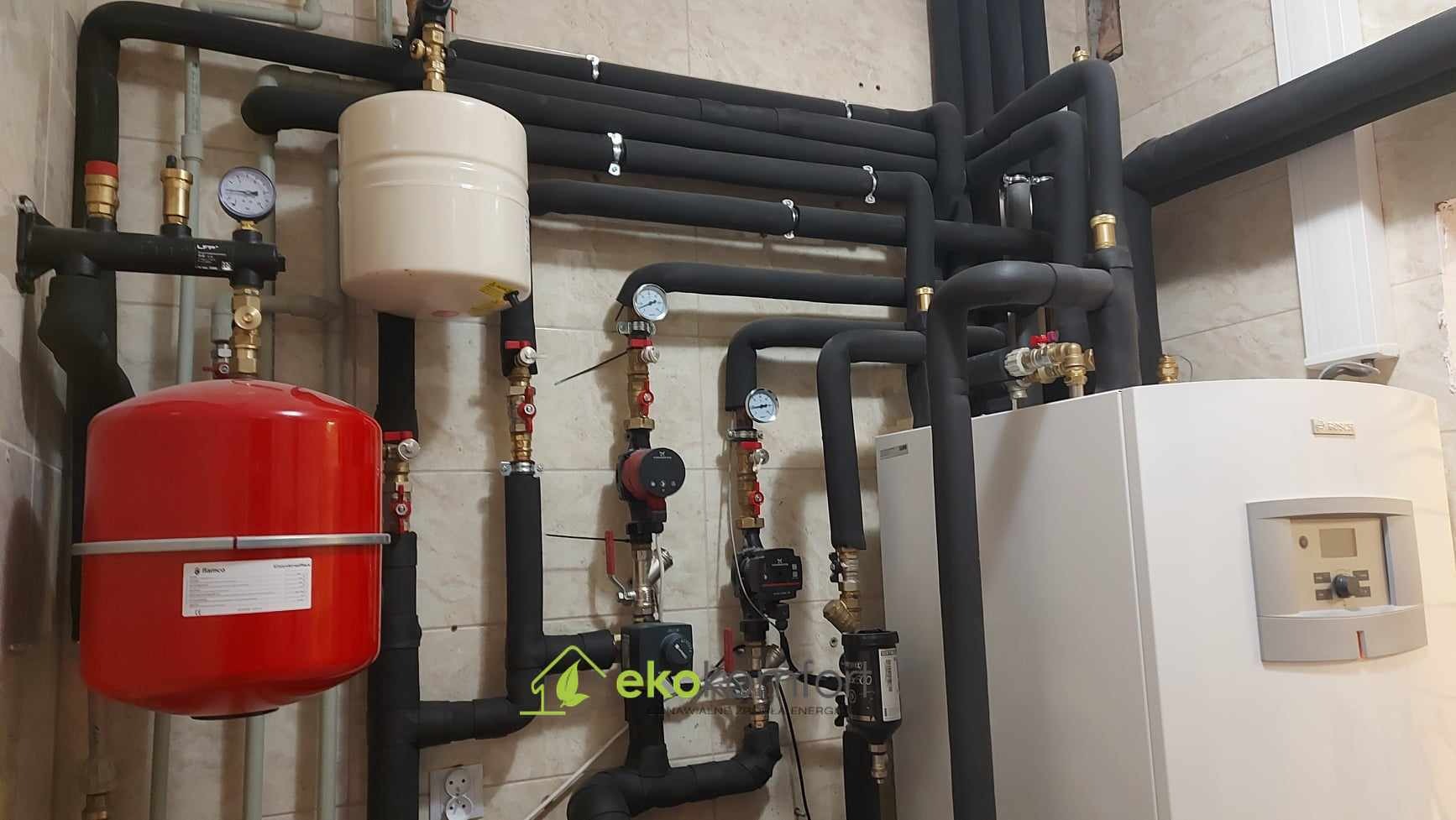 Złożony system rur w izolacji termicznej, manometry, czerwony zbiornik przeponowy, biały zasobnik i inne komponenty instalacji grzewczej na tle beżowych płytek ceramicznych.