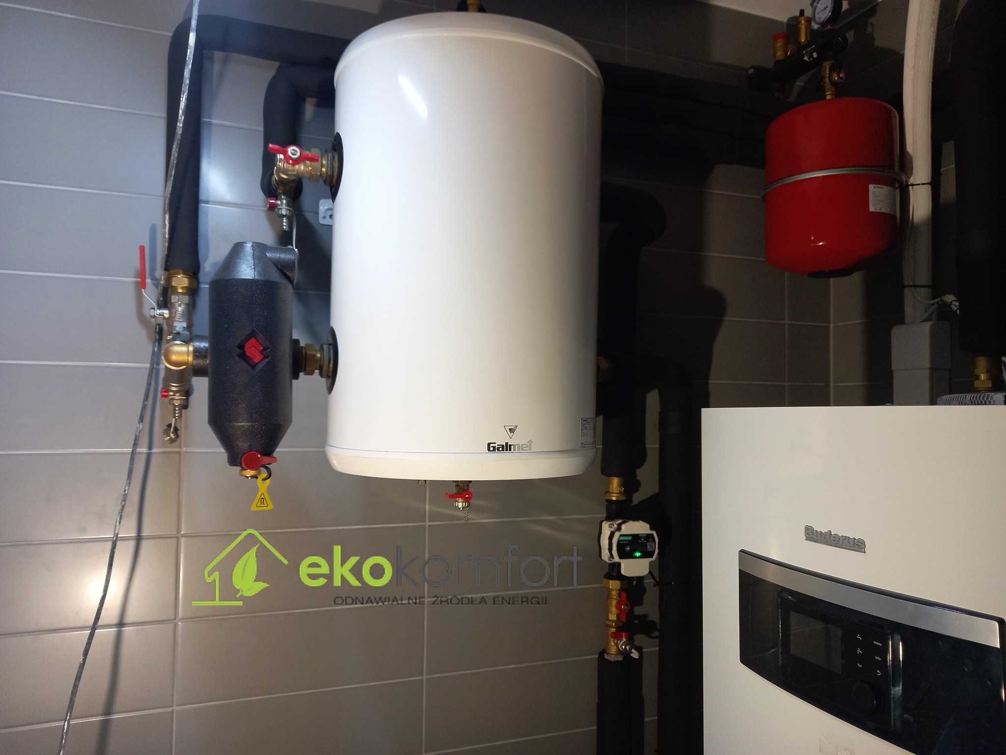 Biały zasobnik ciepłej wody Galmet z czerwonymi zaworami, czarny filtr, czerwony zbiornik wyrównawczy i piec Buderus na tle szarej ściany z płytek, widoczne rury instalacji centralnego ogrzewania...