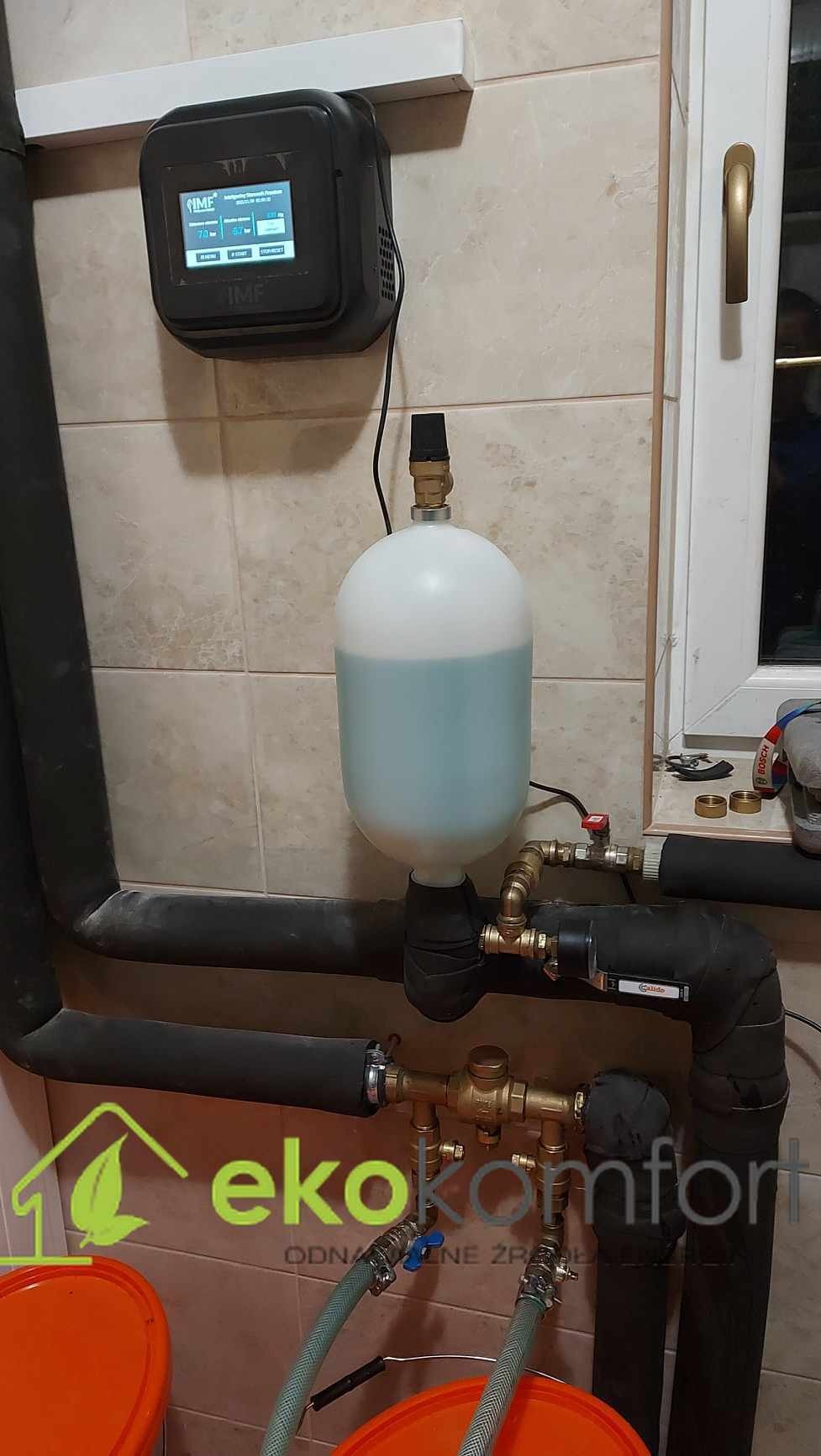 Zamknięty system grzewczy z czarnymi rurami, zbiornikiem wyrównawczym z niebieską cieczą i panelem sterowania IMF na kafelkowanej ścianie, widoczne również dwa pomarańczowe wiadra i fragment okna.
