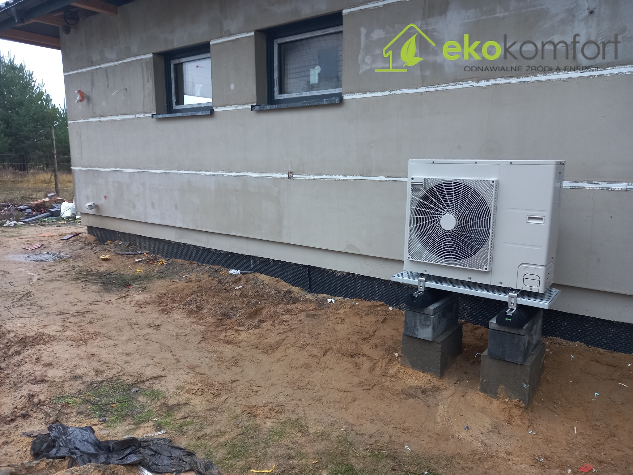 Zewnętrzna jednostka pompy ciepła na betonowych bloczkach przed ocieplonym budynkiem z widocznym logo firmy Eko Komfort na elewacji.