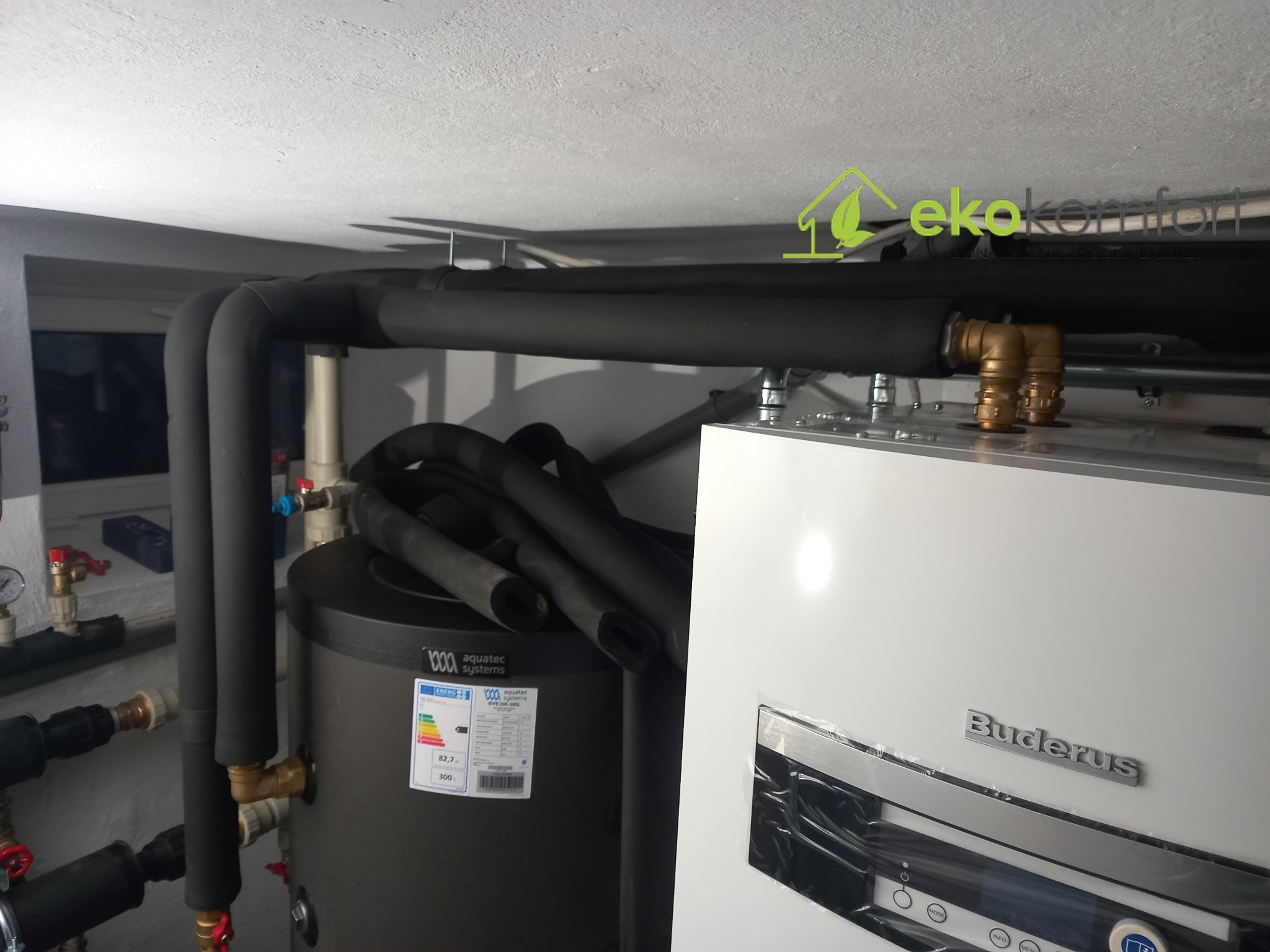 Instalacja grzewcza: biały piec Buderus z podłączonymi czarnymi, izolowanymi rurami, widoczny zbiornik akumulacyjny z etykietą energetyczną, w tle fragment ściany z zaworami.