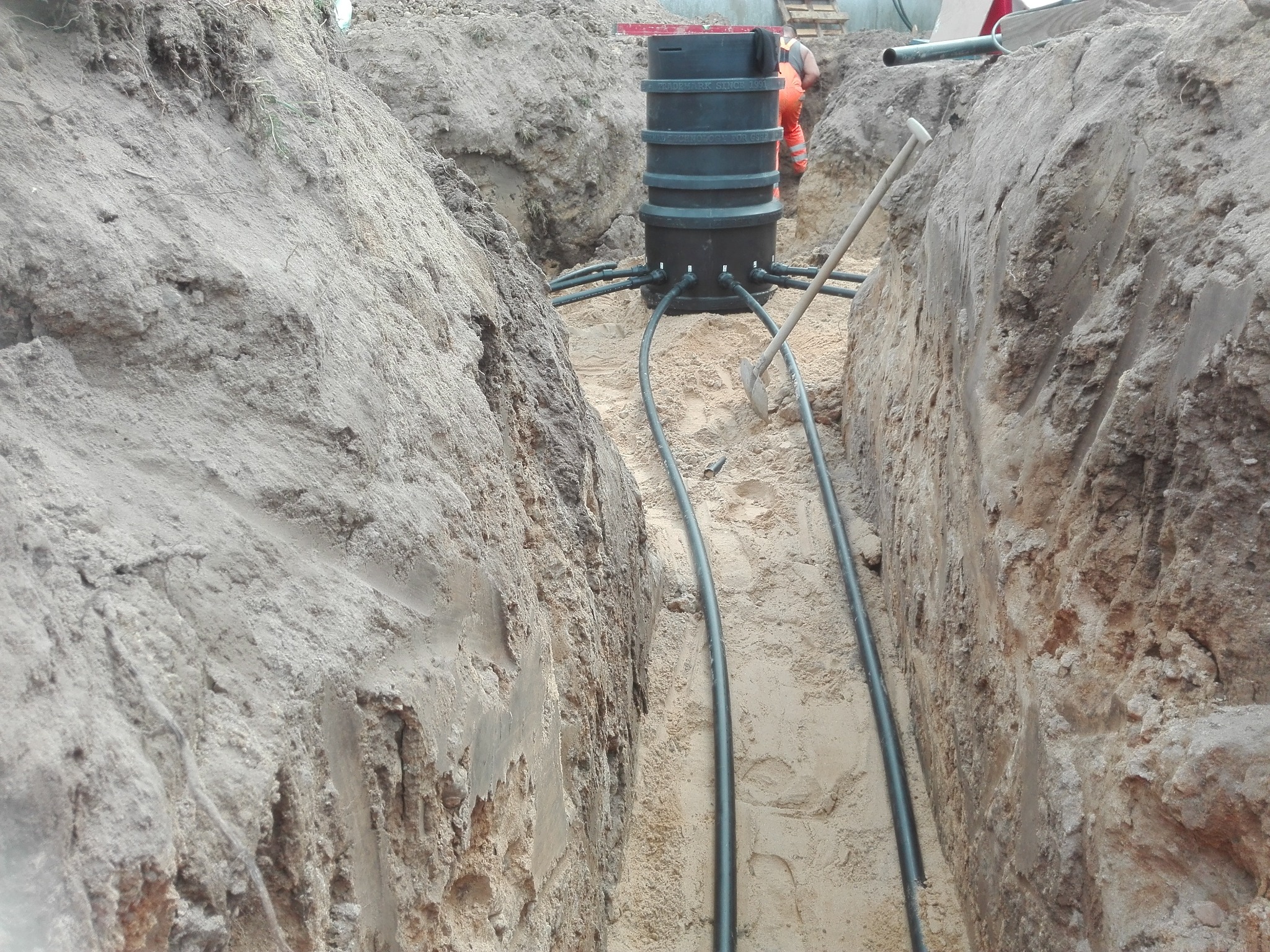 Wykop z piaskowym dnem, w którym umieszczono czarny, cylindryczny zbiornik z podłączonymi rurami; w tle widoczna osoba w pomarańczowym kombinezonie i łopata oparta o zbiornik.