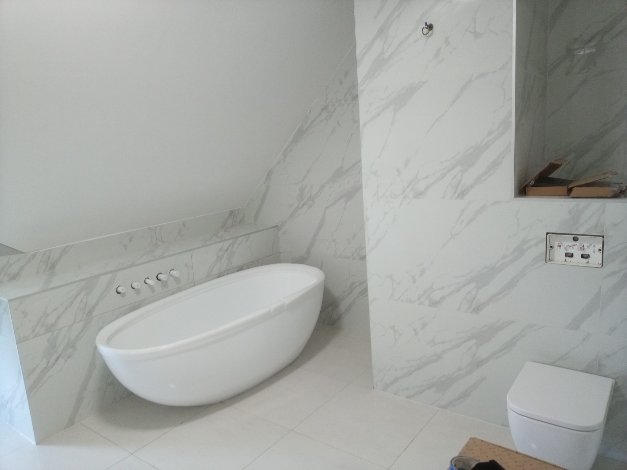 Nowoczesna łazienka w skosie z wanną wolnostojącą i marmurowymi płytkami w trakcie wykańczania, widoczne elementy instalacji i pudełka po płytkach.