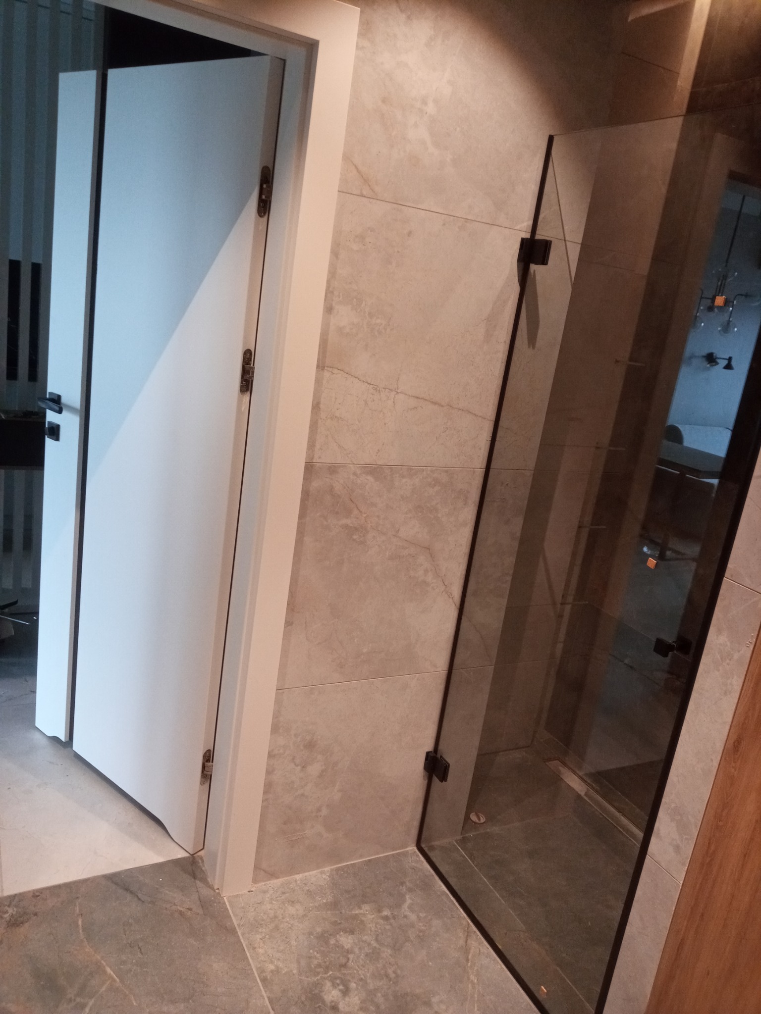 Wykończona łazienka z kabiną prysznicową ze szkła hartowanego i drzwiami w kolorze białym, płytki imitujące marmur na ścianach i podłodze.