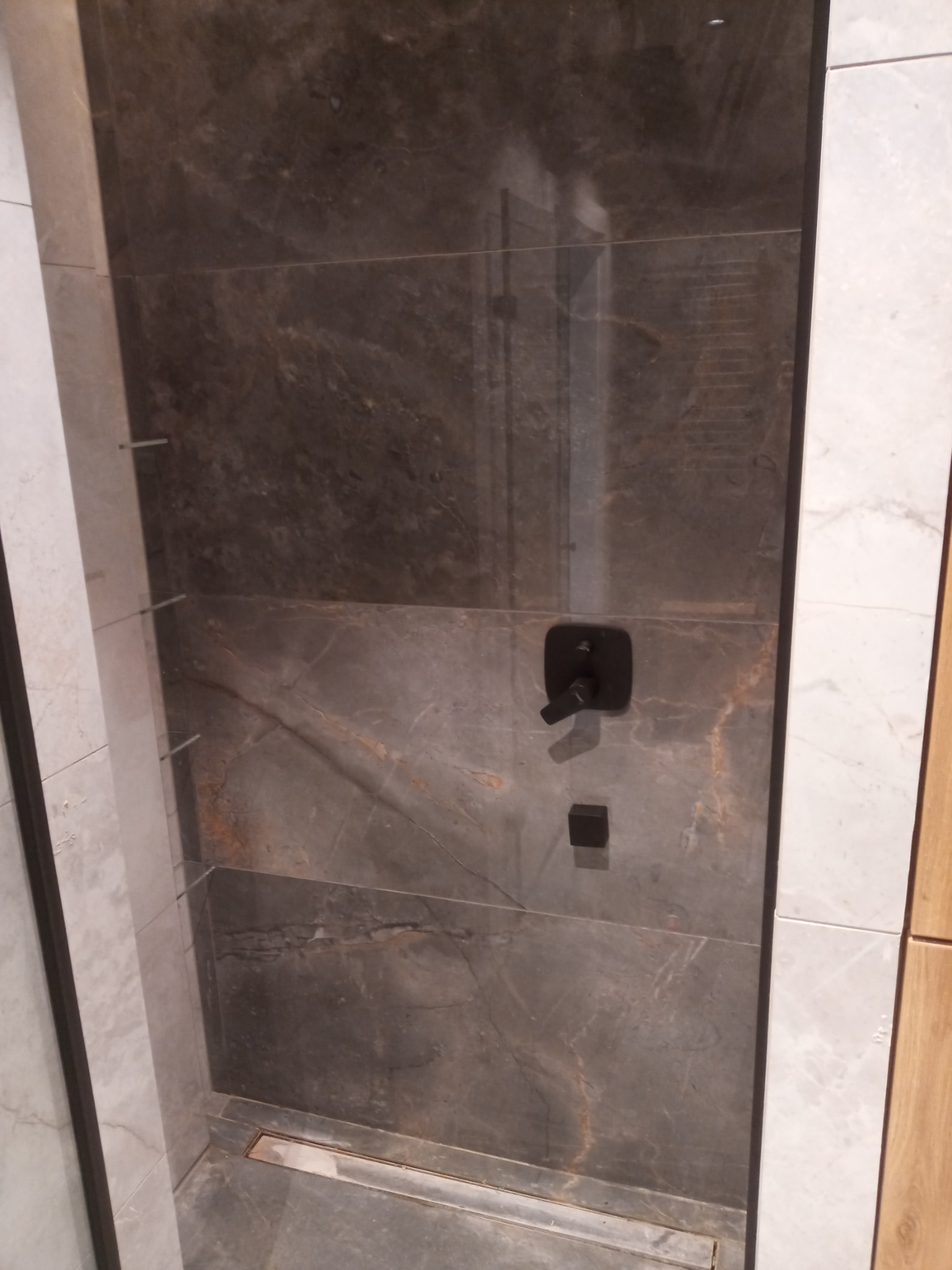 Wykończona kabina prysznicowa z ciemnymi, marmurkowymi płytkami, czarną armaturą i odpływem liniowym w podłodze.