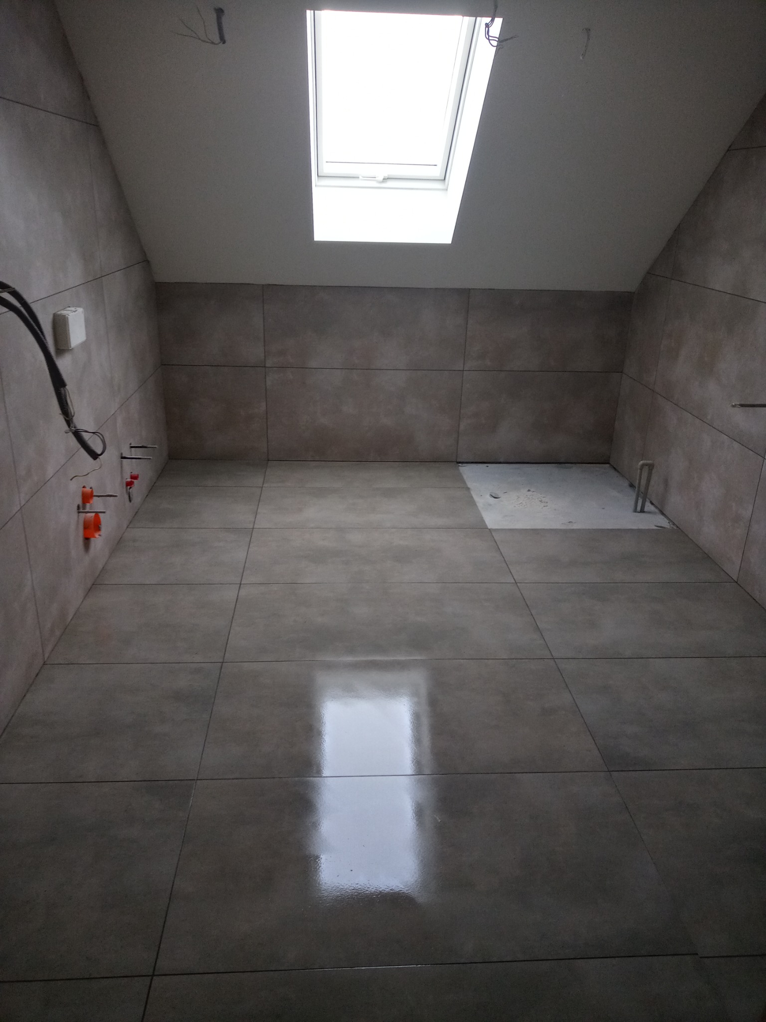 Ukończona łazienka na poddaszu, wyłożona szarymi, matowymi płytkami ceramicznymi, z oknem dachowym i widocznymi instalacjami hydraulicznymi w ścianie.
