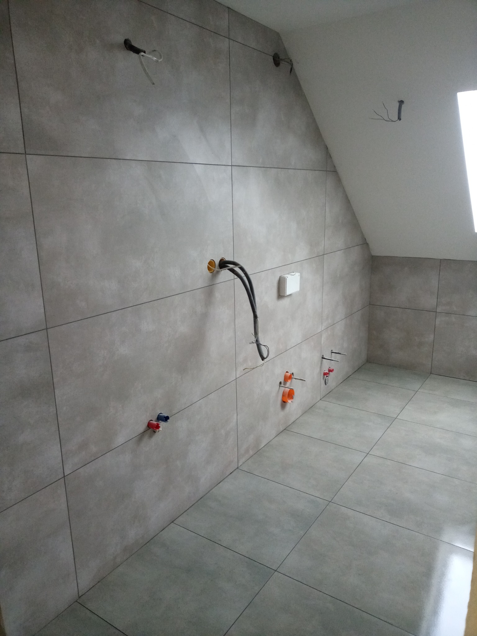 Wnętrze łazienki w trakcie remontu, ściany i podłoga wyłożone szarymi płytkami imitującymi kamień, widoczne wyprowadzenia instalacji wodnej i elektrycznej na ścianach, sufit ze skosem.