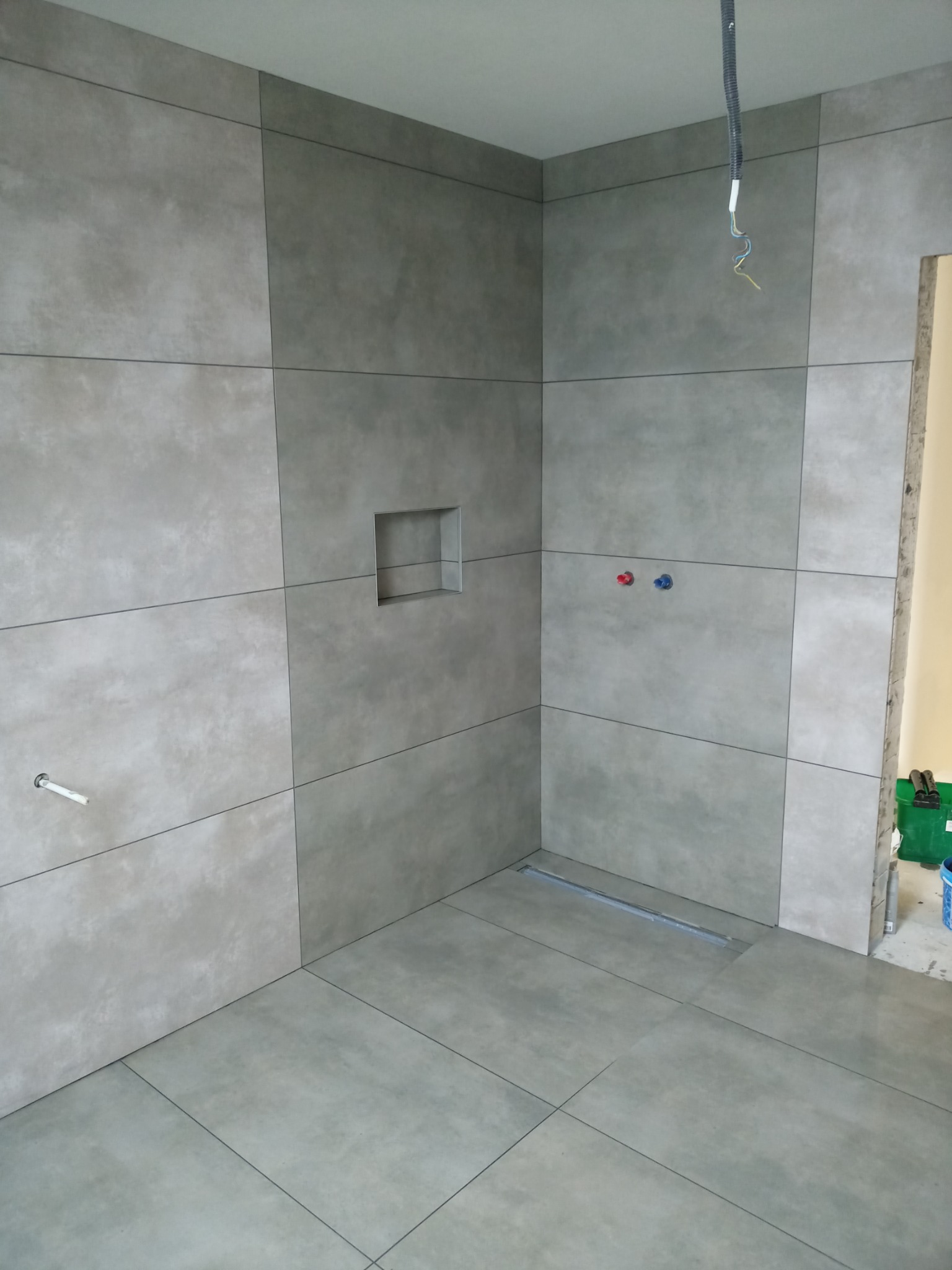 Wyłożona szarymi, matowymi płytkami łazienka w trakcie remontu; widoczny odpływ liniowy, wystające kable i niezamaskowane przyłącza wody.