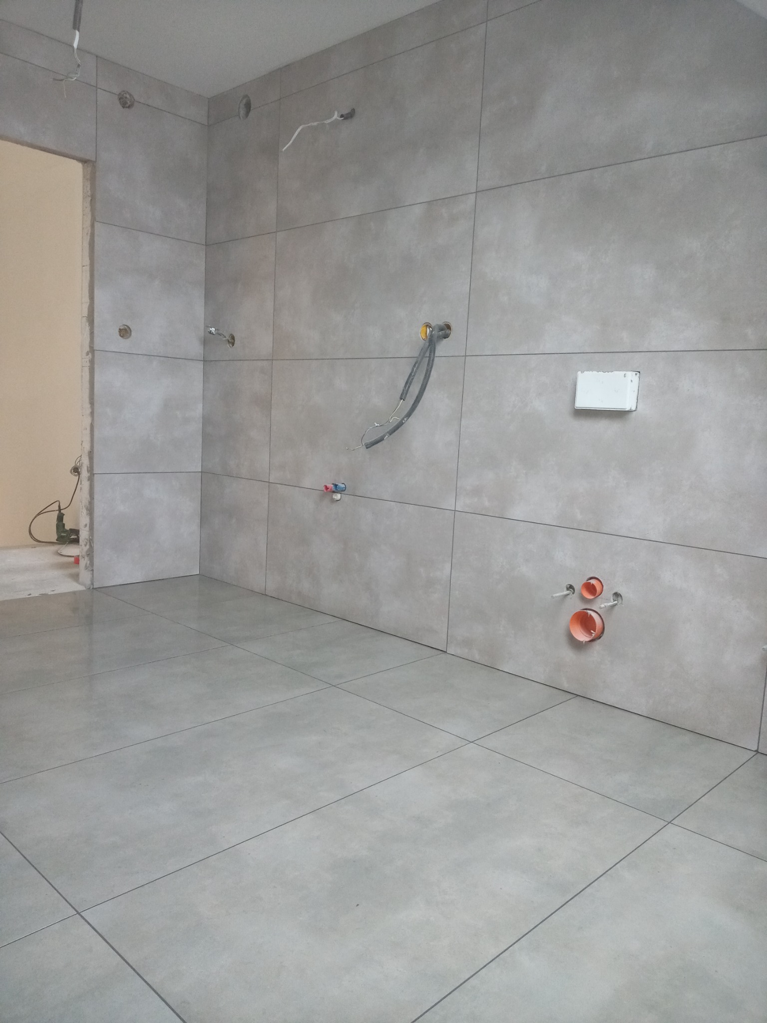 Wyłożona szarymi płytkami łazienka w trakcie remontu, widoczne wystające rury i kable, brak białej armatury.