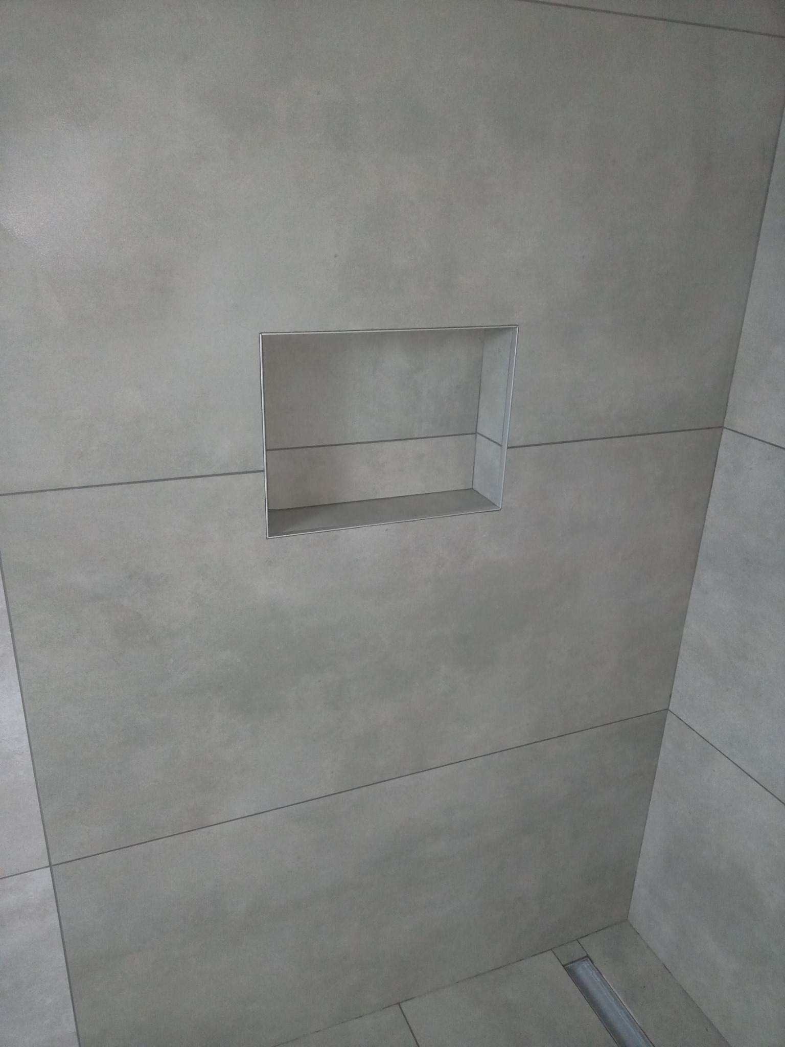 Ściana wyłożona szarymi płytkami z wnęką w kształcie prostokąta z metalowym obramowaniem, widoczny odpływ liniowy w podłodze.