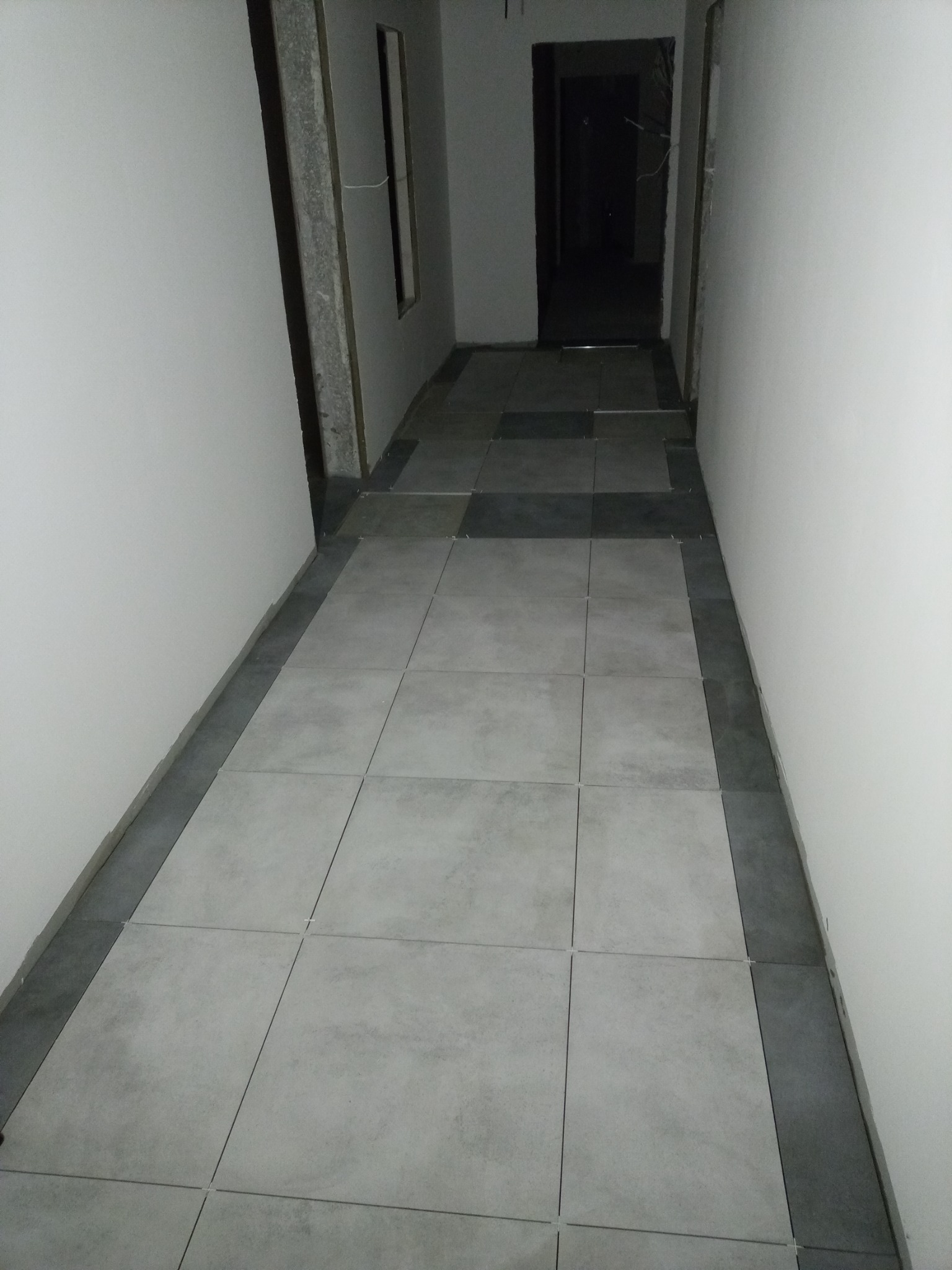 Korytarz w trakcie układania jasnoszarych płytek podłogowych z ciemnoszarą obwódką przy ścianach; widoczne krzyżyki dystansowe.