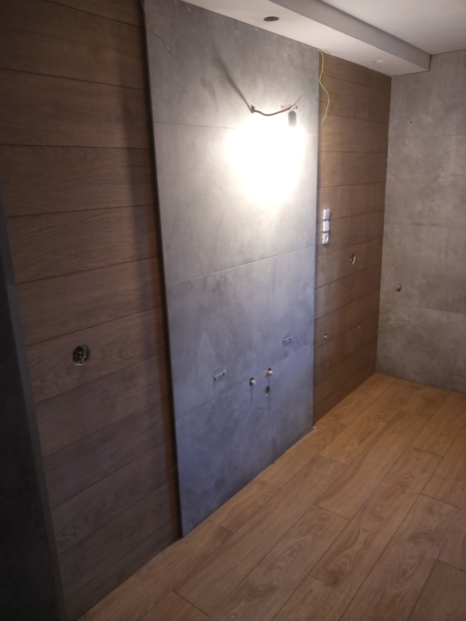 Ściana w trakcie wykańczania z płytek imitujących drewno i szarej płyty, widoczne przewody elektryczne i instalacja hydrauliczna.