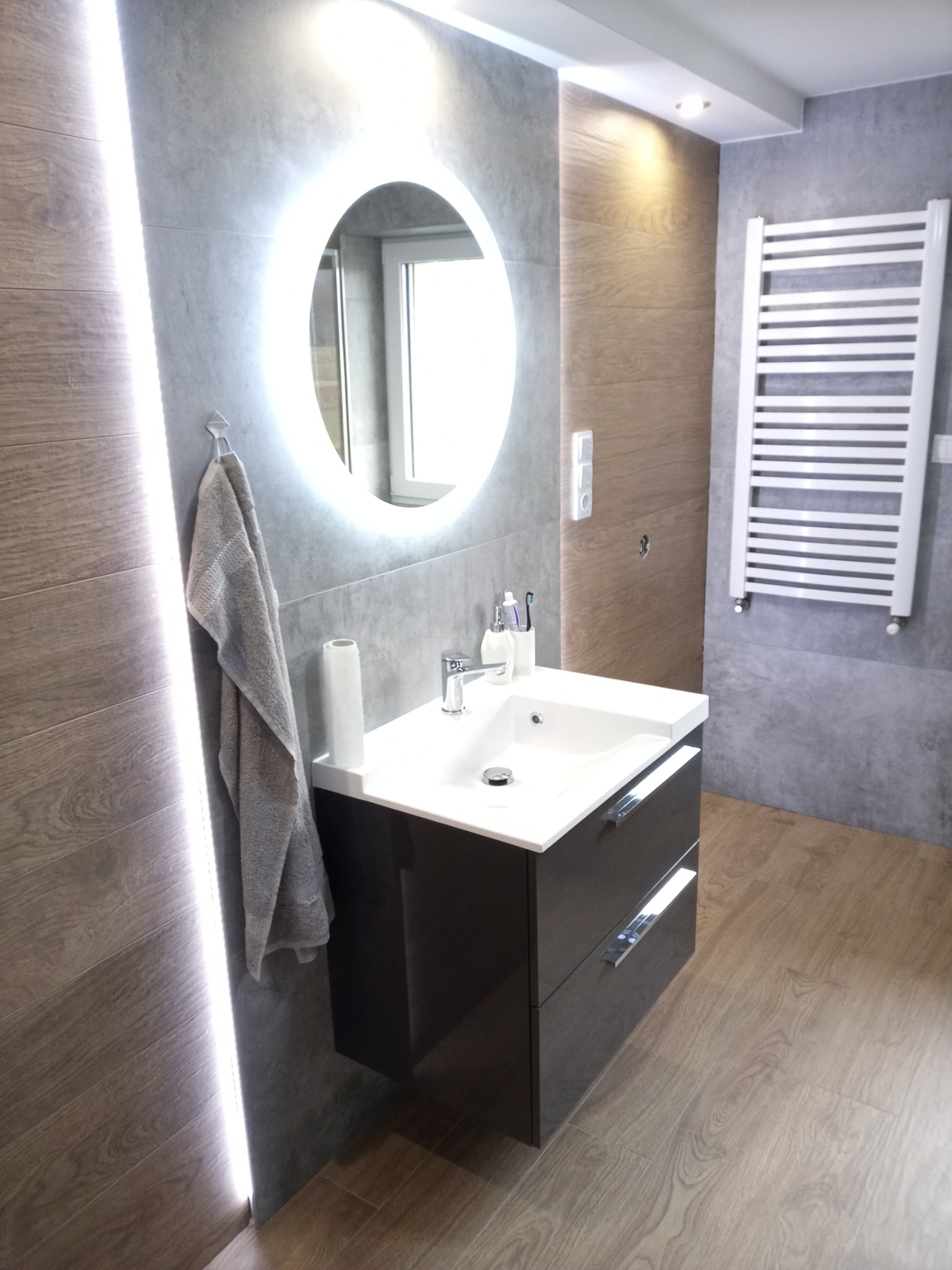 Nowoczesna łazienka z okrągłym lustrem podświetlanym LED, szara glazura imitująca drewno na ścianach i podłodze, oraz biały grzejnik drabinkowy.