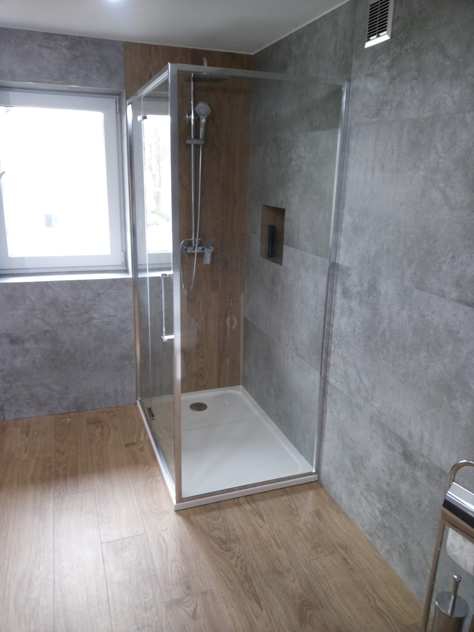 Nowoczesna kabina prysznicowa z przezroczystego szkła, biały brodzik, szare płytki imitujące beton i drewnopodobne panele na ścianie, widoczna wentylacja sufitowa.