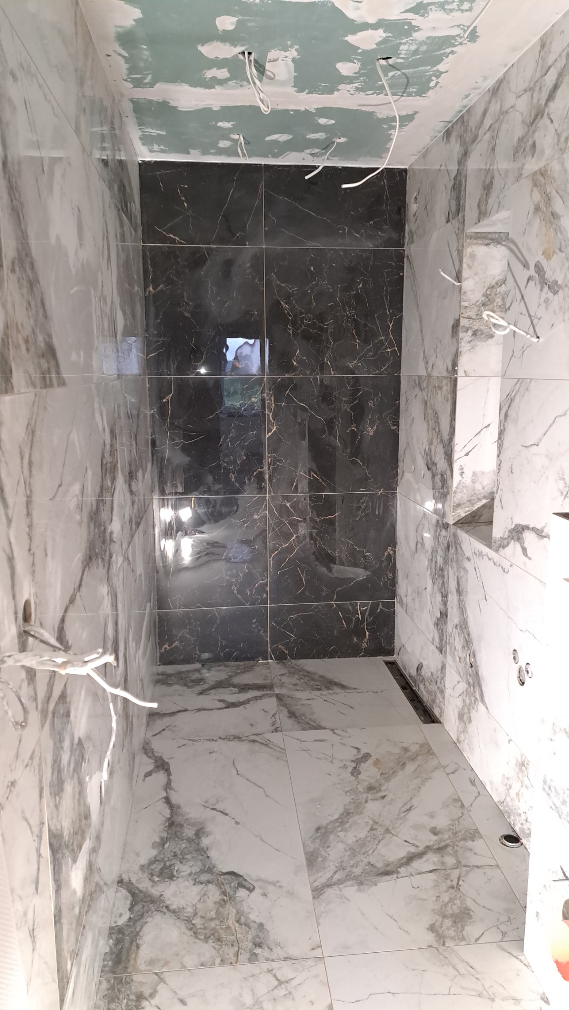 Wnętrze łazienki w trakcie remontu z wyłożonymi marmurowymi płytkami na ścianach i podłodze, z ciemną wstawką na jednej ze ścian oraz widocznymi przewodami elektrycznymi wystającymi z sufitu.