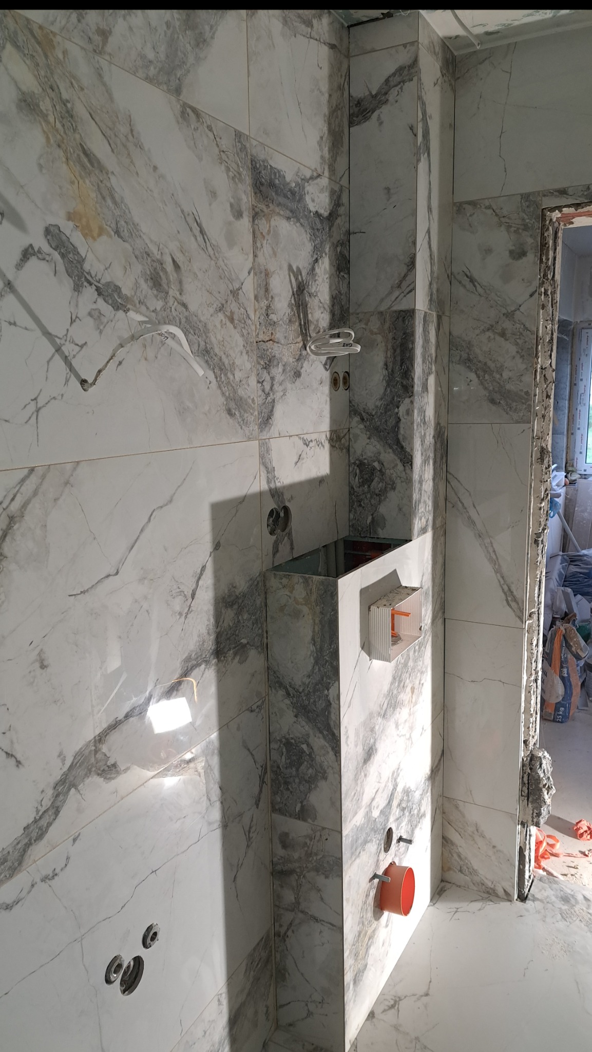 Łazienka w trakcie remontu: ściany i obudowa stelaża WC wyłożone białymi płytkami imitującymi marmur z szarymi żyłami, widoczne otwory na armaturę i rury kanalizacyjne, surowa ościeżnica drzwi.
