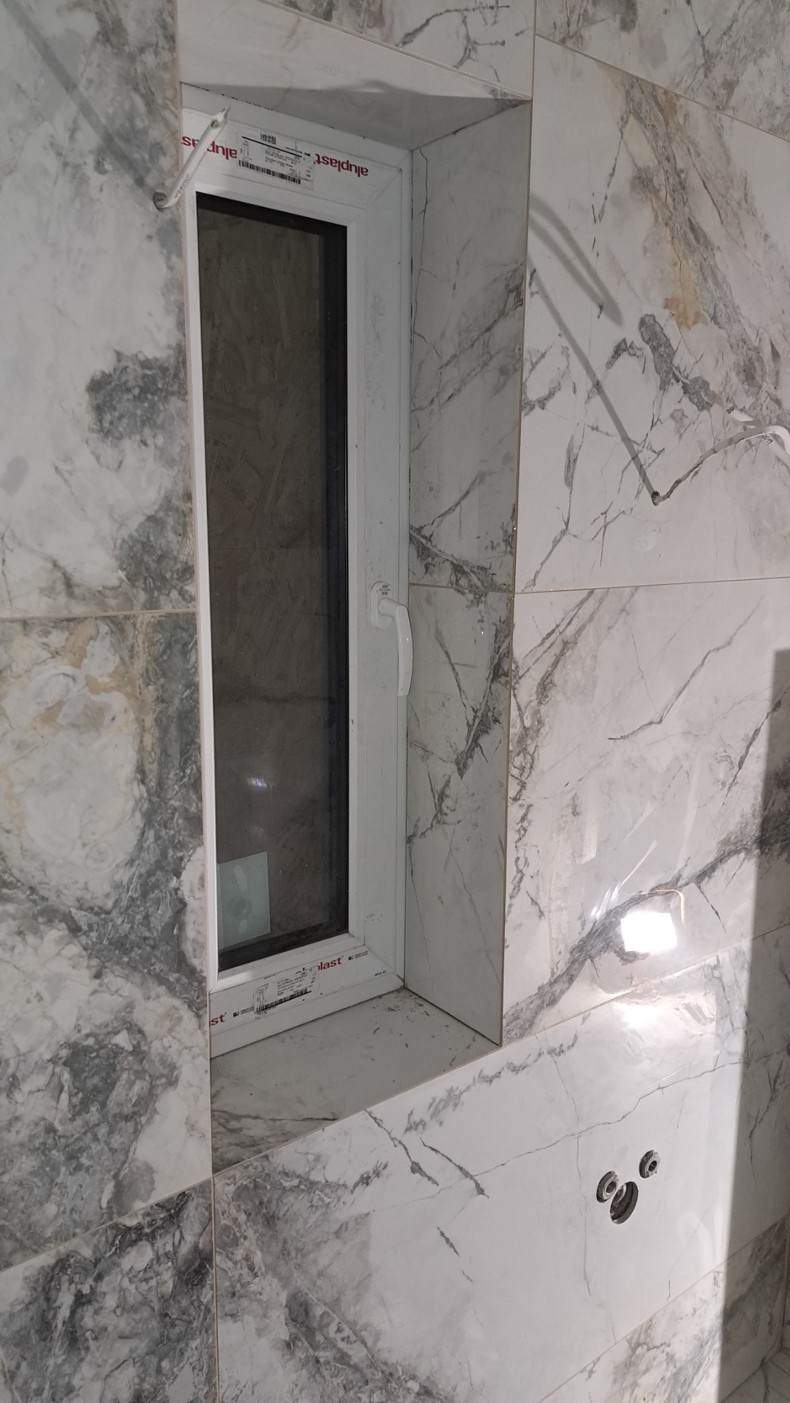 Wykończona wnęka okienna w łazience z płytkami imitującymi marmur, widoczne otwory na baterię.