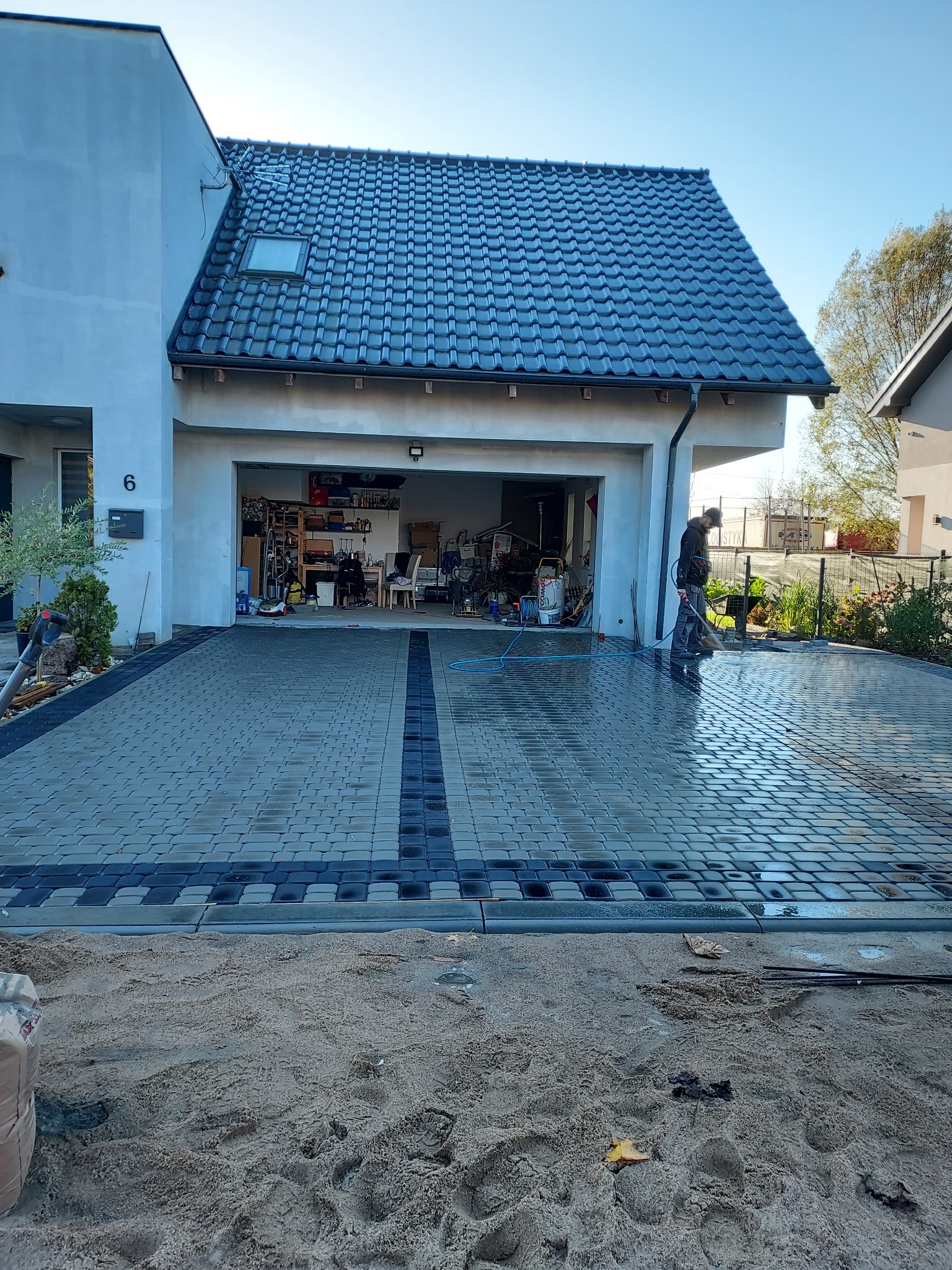 Świeżo ułożona, mokra kostka brukowa w odcieniach szarości z ciemniejszym pasem pośrodku, przed nowoczesnym domem z otwartym garażem, pracownik myje powierzchnię wodą pod ciśnieniem, widoczny...