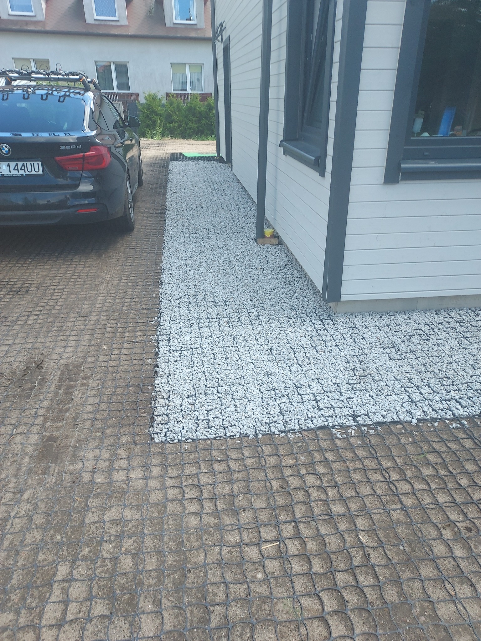 Niedawno ukończony podjazd z plastikowej kratki wypełnionej drobnym, białym kruszywem, sąsiadujący z podjazdem z kratki wypełnionej ziemią, obok zaparkowany czarny samochód BMW.