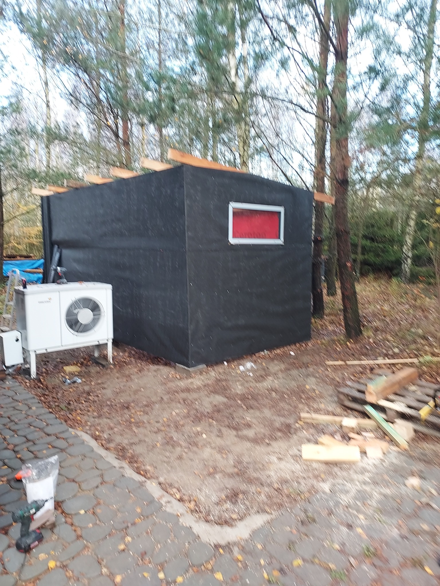 Czarna, tymczasowa konstrukcja z czerwonym obramowaniem okna, otoczona drzewami i sprzętem budowlanym, w tym pompą ciepła i narzędziami na kostce brukowej.