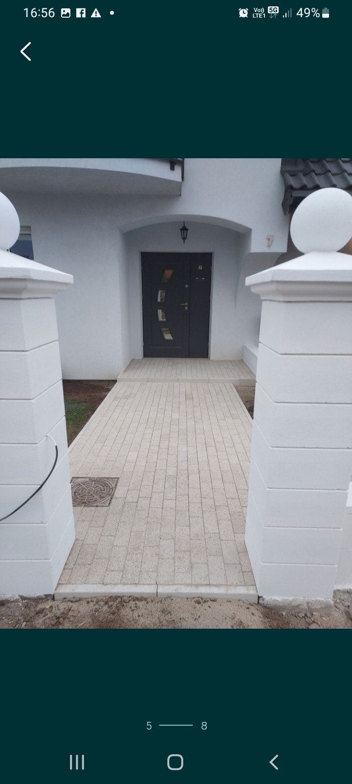 Wejście do domu z szarą kostką brukową, widoczne dwa białe słupki z kulami na szczycie, szare drzwi wejściowe i numer domu nad drzwiami.