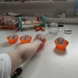 Bielskie Centrum Logopedyczne Aneta Franczuk - Dziecięca ręka sięga do pomarańczowej papilotki z literą 'i', obok papilotki z literami 'A' i 'U' na białym stole z zabawkami w tle.