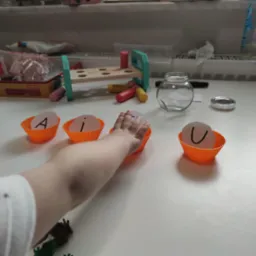 Dziecięca ręka sięga do pomarańczowej papilotki z literą 'i', obok papilotki z literami 'A' i 'U' na białym stole z zabawkami w tle.