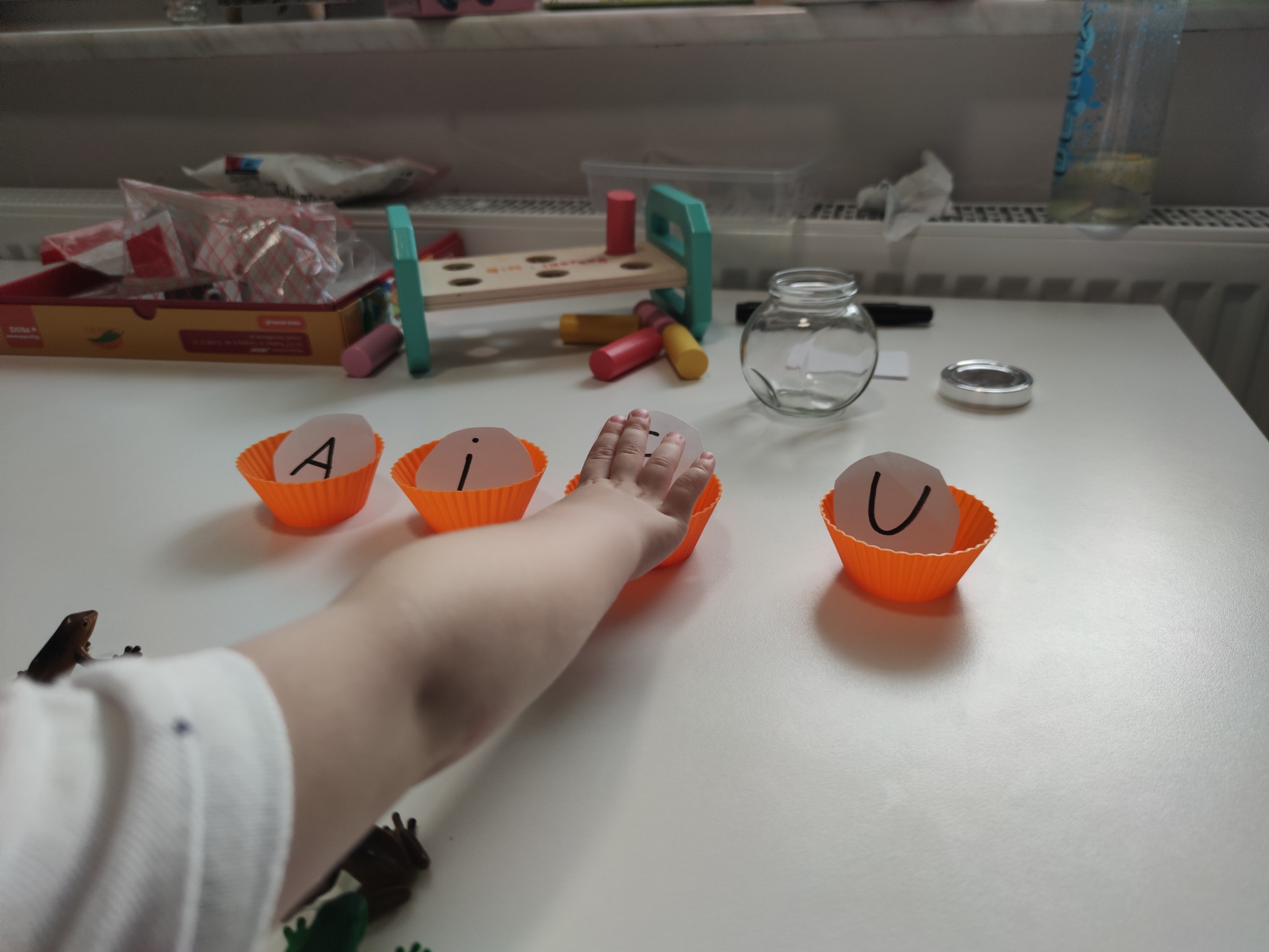 Dziecięca ręka sięga do pomarańczowej papilotki z literą 'i', obok papilotki z literami 'A' i 'U' na białym stole z zabawkami w tle.