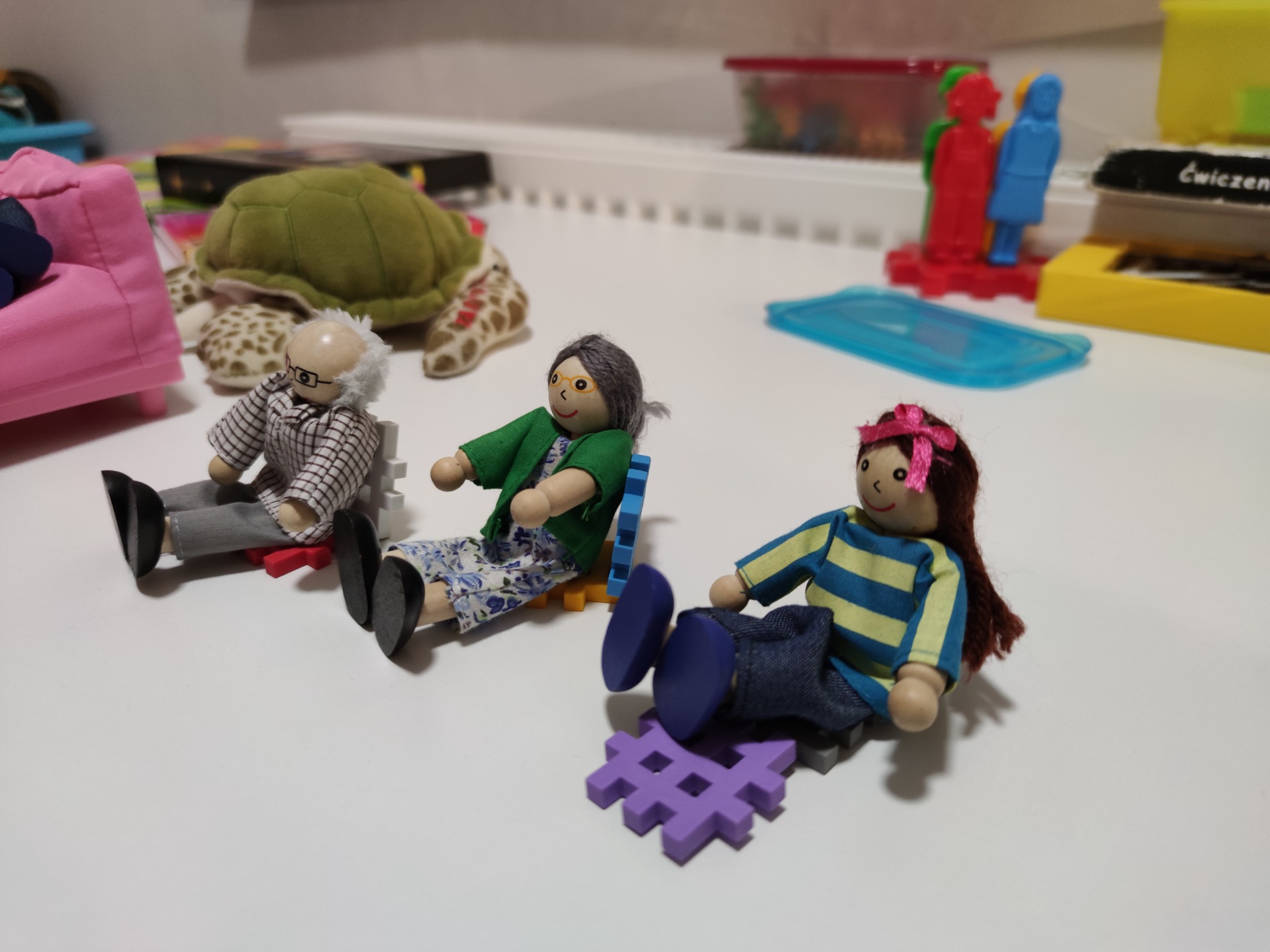 Trzy drewniane figurki przedstawiające osoby siedzące na małych krzesełkach z klocków, w tle pluszowa żółw i zabawki edukacyjne.