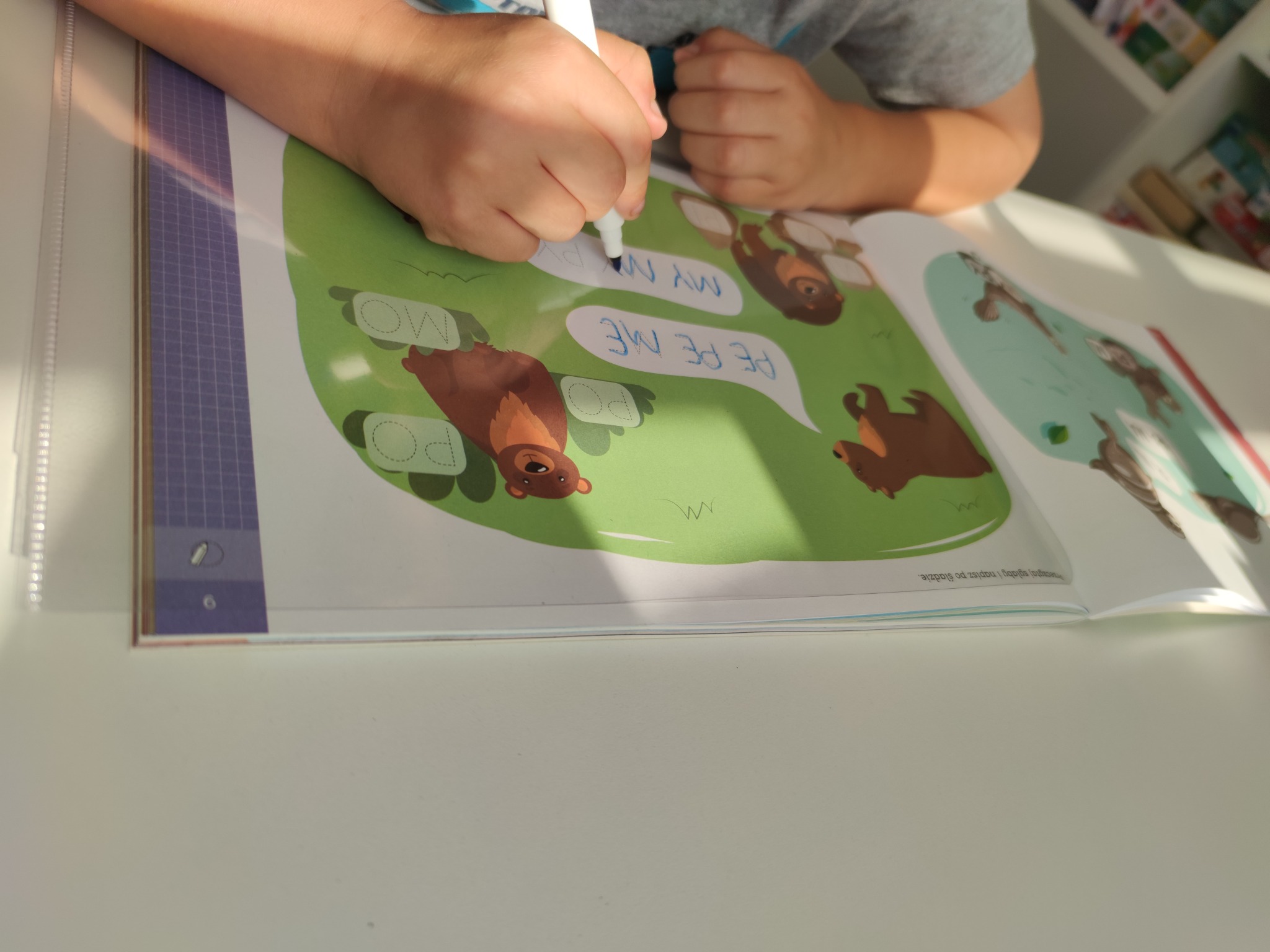 Dziecko ćwiczy pisanie po śladzie w książce z obrazkami niedźwiedzi, używając białego flamastra na stole w pobliżu regału z książkami.