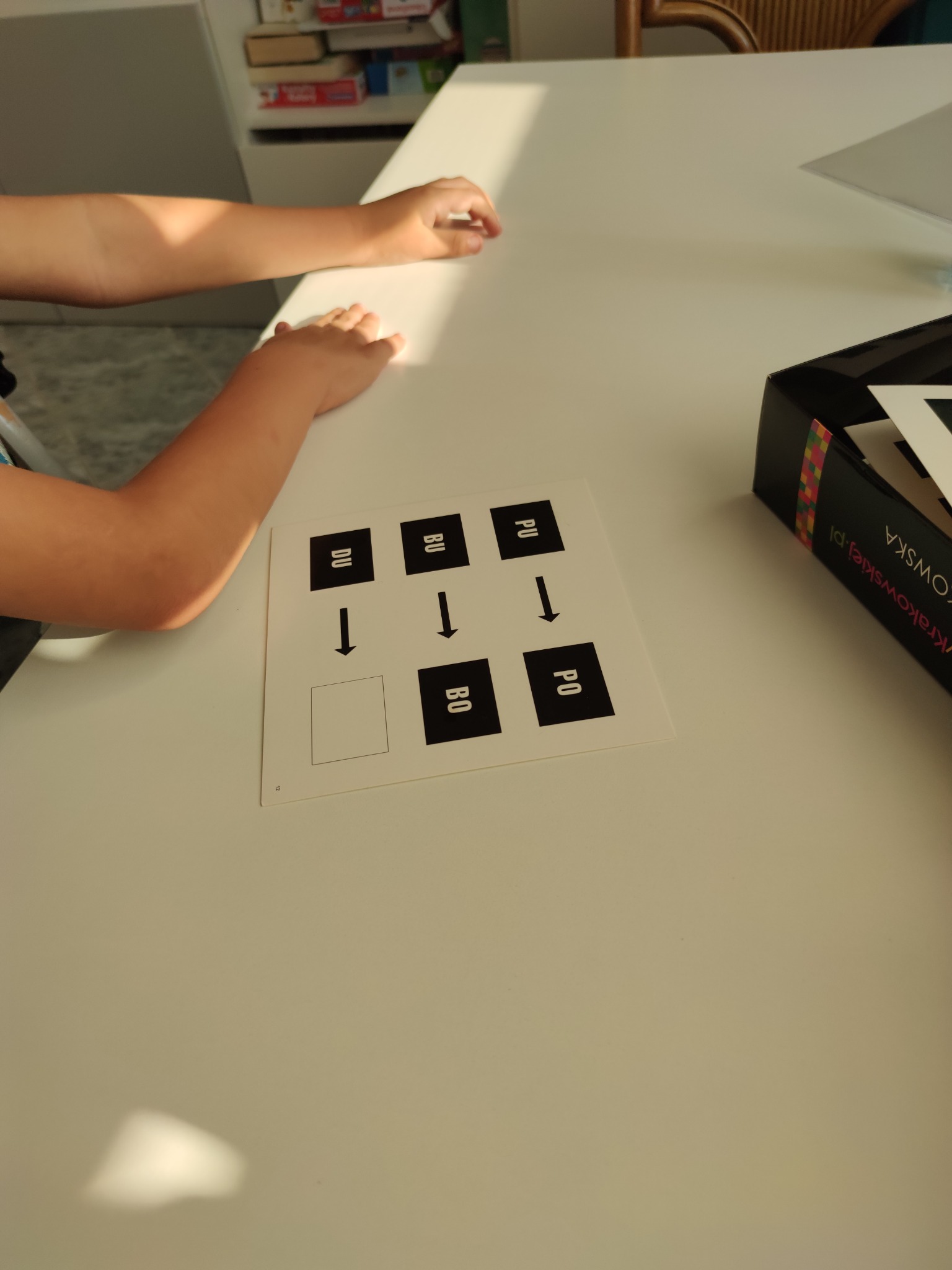 Dziecko siedzi przy stole z kartką z ćwiczeniami, na której znajdują się strzałki i sylaby w kwadratach, obok leży pudełko z grą.