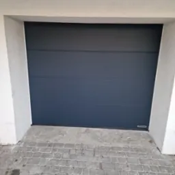 Zamknięta, antracytowa brama garażowa segmentowa z poziomymi przetłoczeniami, widoczny fragment kostki brukowej przed wjazdem i logo producenta w prawym dolnym rogu.