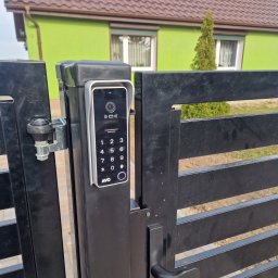 NA PILOTA ŁUKASZ ZAJĄKAŁA - Nowoczesny wideodomofon z klawiaturą numeryczną zamontowany na czarnej, poziomej bramie wjazdowej; w tle zielony dom z brązowym dachem.