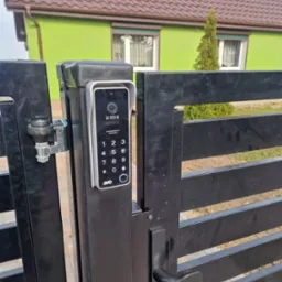 Nowoczesny wideodomofon z klawiaturą numeryczną zamontowany na czarnej, poziomej bramie wjazdowej; w tle zielony dom z brązowym dachem.