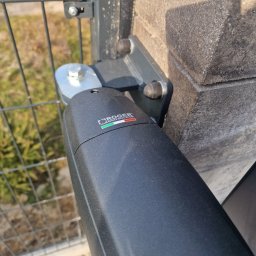 NA PILOTA ŁUKASZ ZAJĄKAŁA - Szczegół montażu napędu bramy na słupku ogrodzeniowym, widoczne mocowanie oraz logo producenta.
