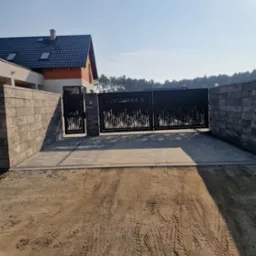 Nowoczesna brama wjazdowa z metaloplastyki z motywem roślinnym, zamontowana na posesji z kostki brukowej i szarego muru, widoczne ślady opon na piasku.