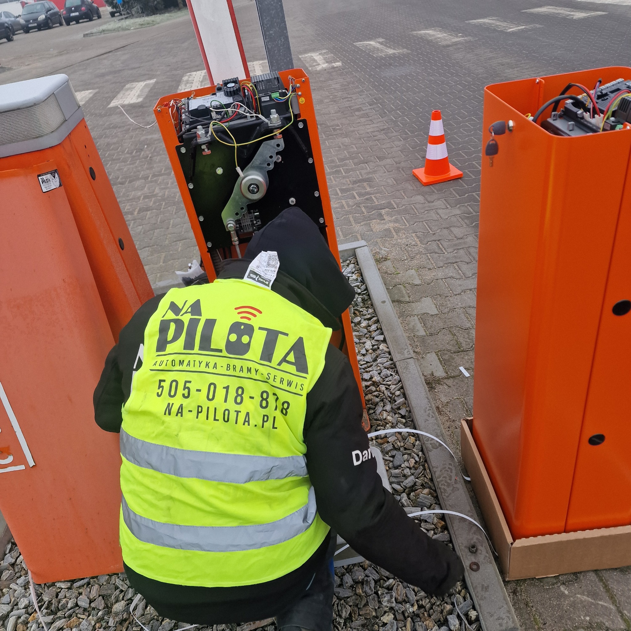 Serwisant w żółtej kamizelce z logo firmy naprawia pomarańczową obudowę szlabanu automatycznego na parkingu, widoczne otwarte wnętrze z elektroniką i mechanizmami.