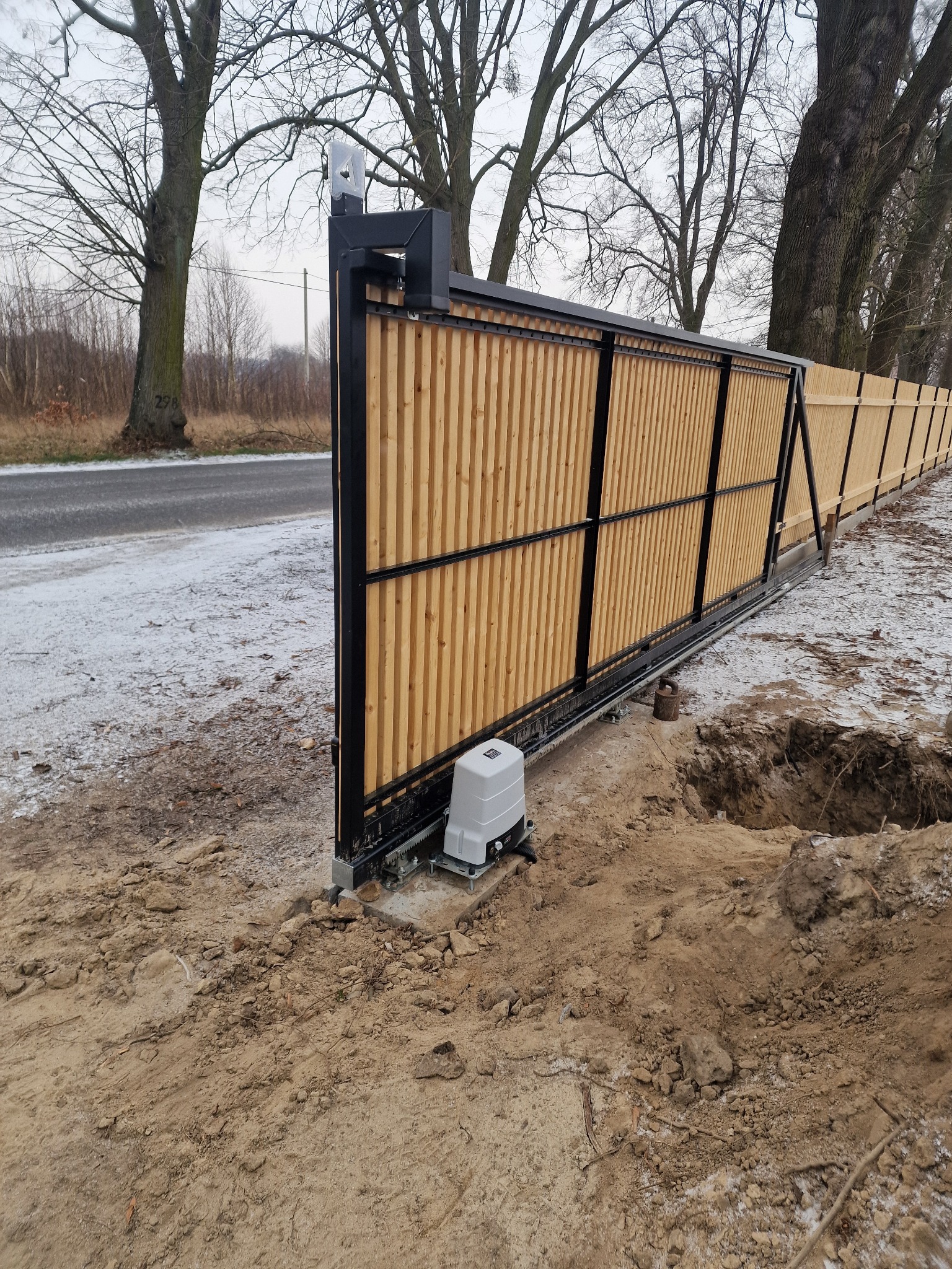 Nowa brama przesuwna z drewnianymi lamelami i automatyką, widoczny silnik napędu, teren po montażu wymaga uporządkowania.