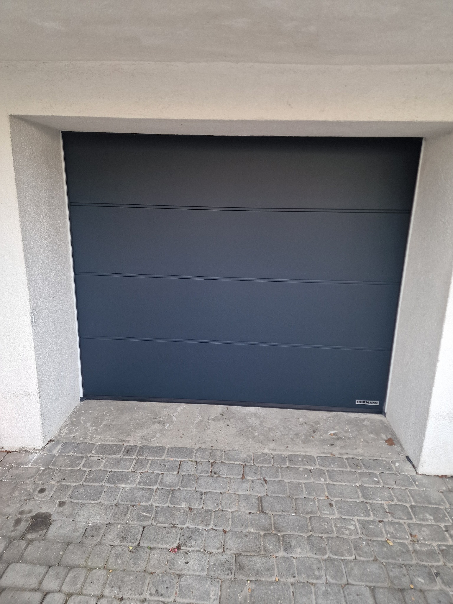 Zamknięta, antracytowa brama garażowa segmentowa z poziomymi przetłoczeniami, widoczny fragment kostki brukowej przed wjazdem i logo producenta w prawym dolnym rogu.