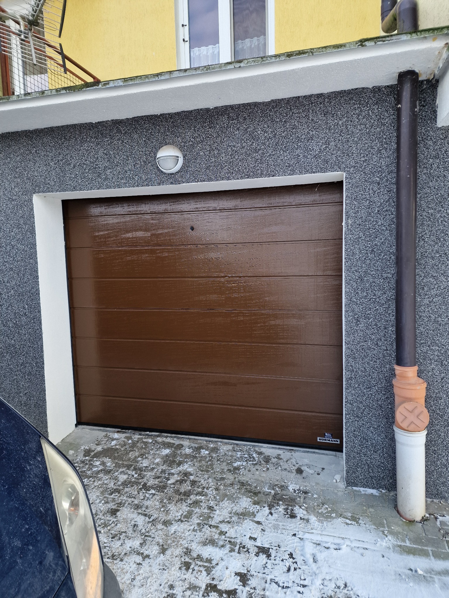 Brązowa brama garażowa segmentowa Hormann z poziomymi przetłoczeniami, widoczna z boku, z fragmentem zaparkowanego samochodu i zaśnieżonym podjazdem.