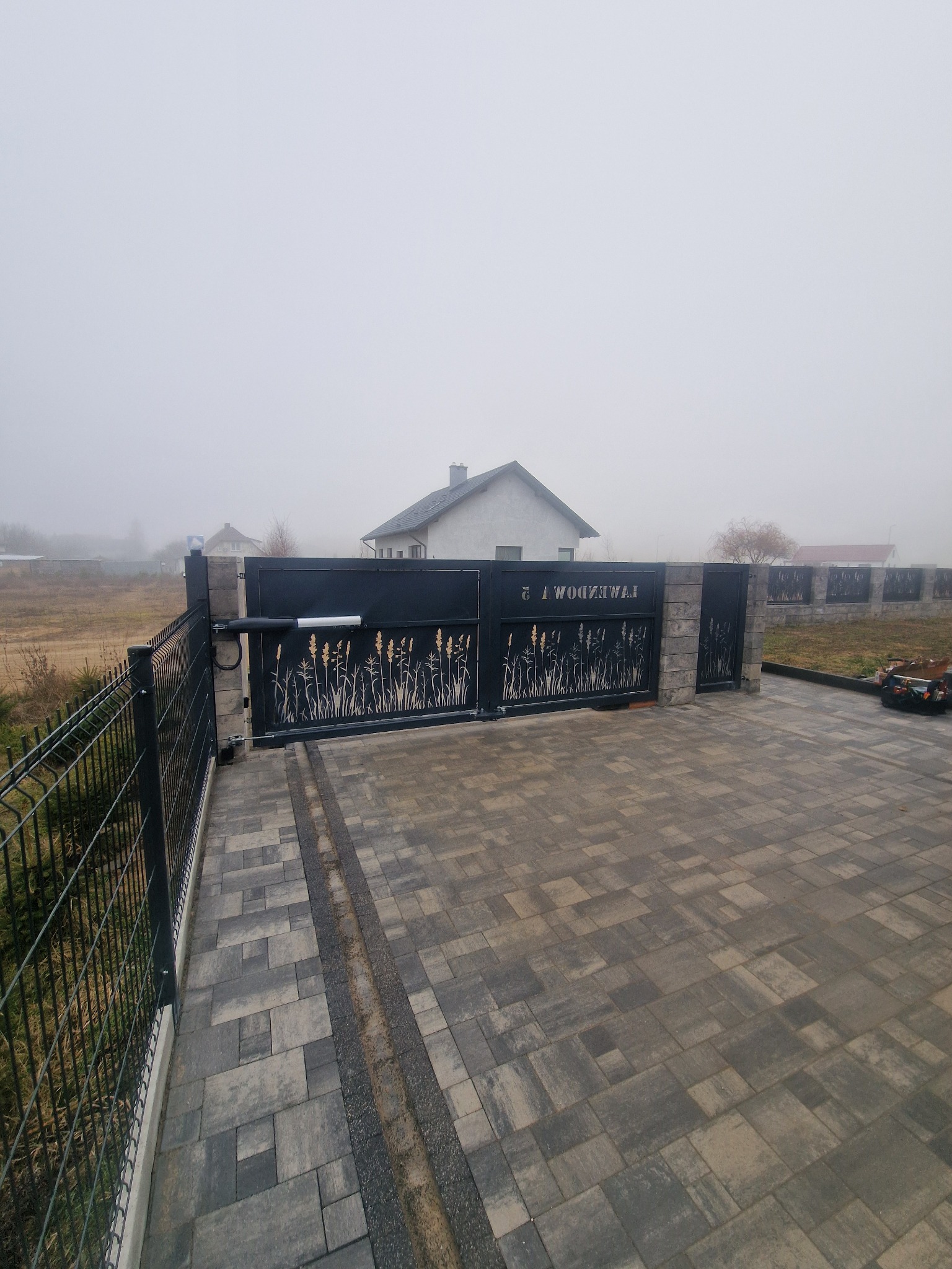 Automatyczna brama wjazdowa z motywem roślinnym, zamontowana na posesji z kostką brukową w mglisty dzień.